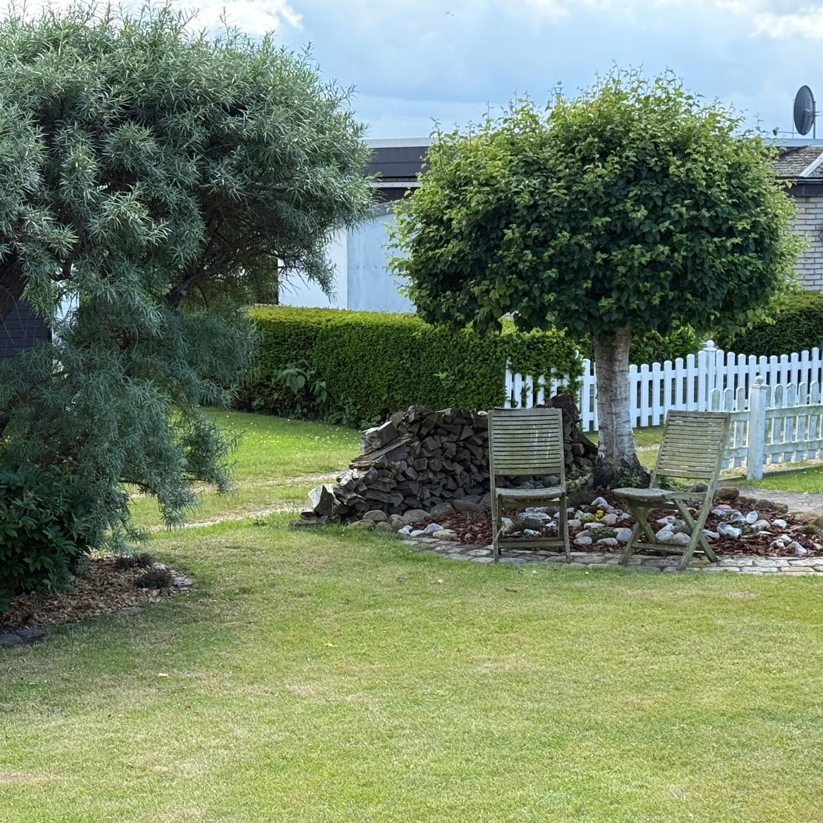 Garten mit zwei Holzstühlen, Baum, Holzstapel und weißer Zaun.