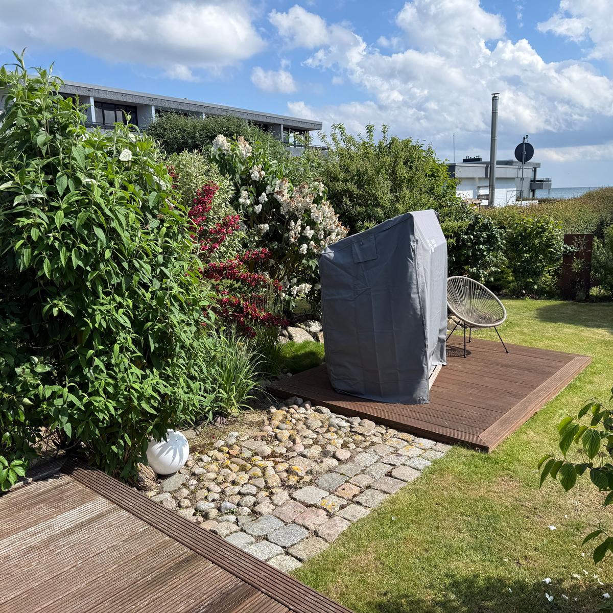 Garten mit Holzterrasse, Beete mit Blumen und einem gedeckten Grill.