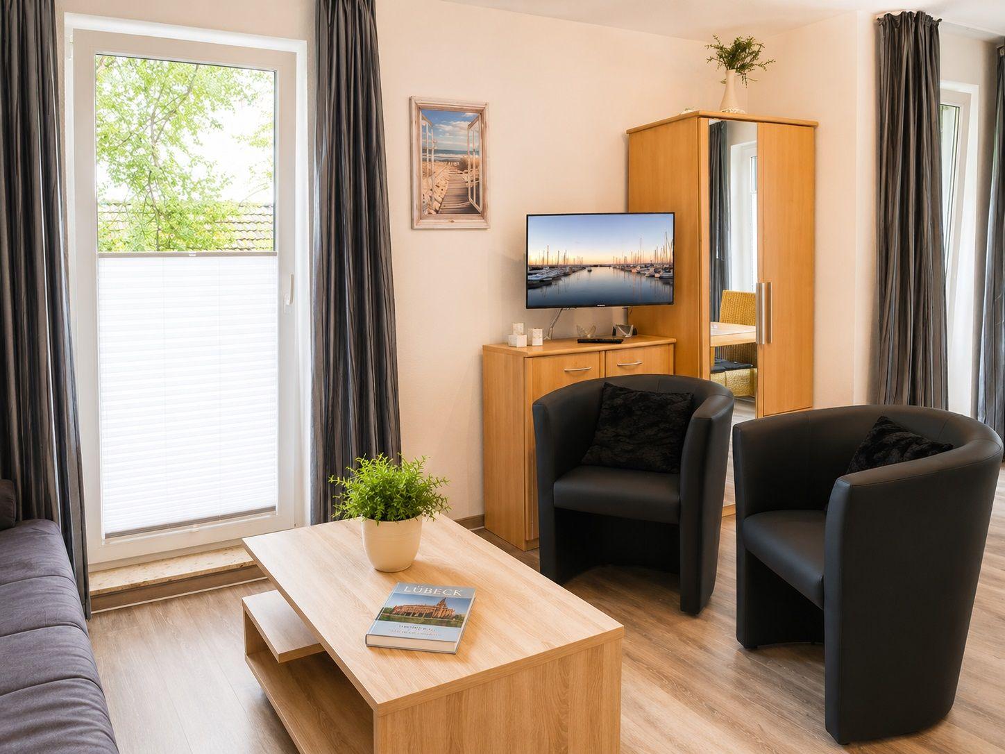 Wohnzimmer mit zwei schwarzen Sesseln, Holztisch und Fernseher. Fenster mit Blick auf Grün.