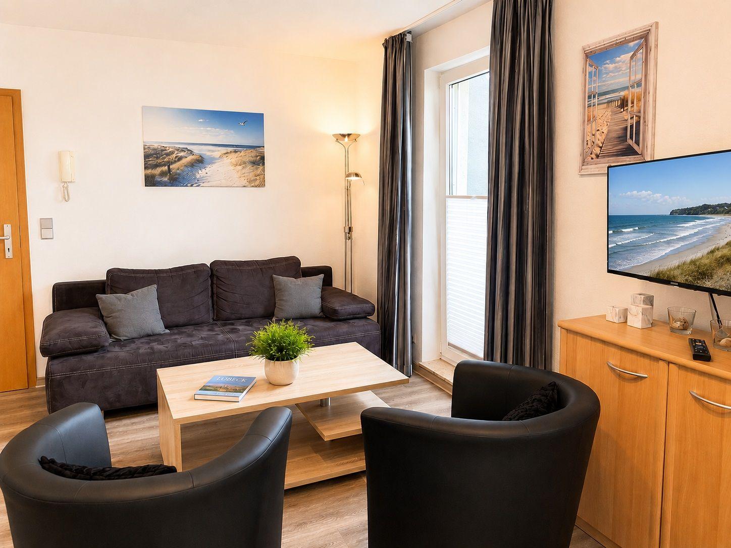 Wohnzimmer mit Sofa, zwei Sesseln, Tisch und TV. Wandbild und Fenster mit Vorhängen.