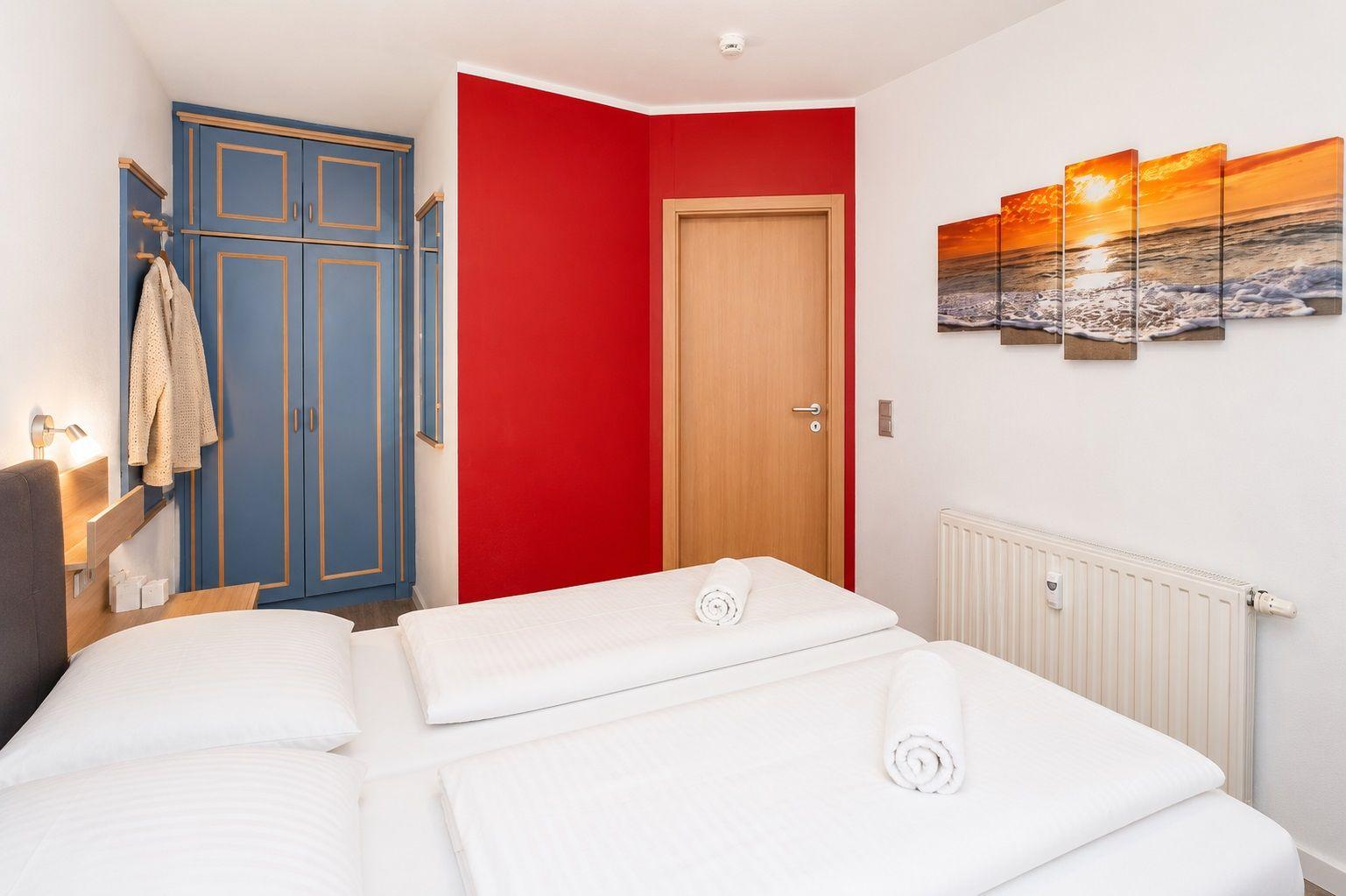Schlafzimmer mit Doppelbett, blauem Schrank und roter Wand. Wandbild zeigt Sonnenuntergang.