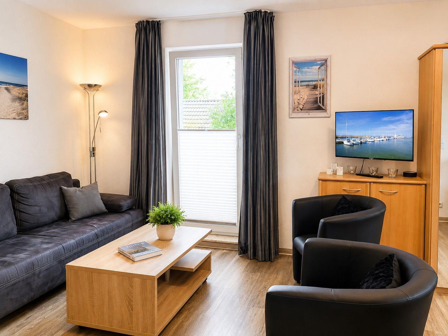 Wohnzimmer mit Sofa, zwei Sesseln, Couchtisch und TV. Fenster mit Jalousie und Vorhängen.