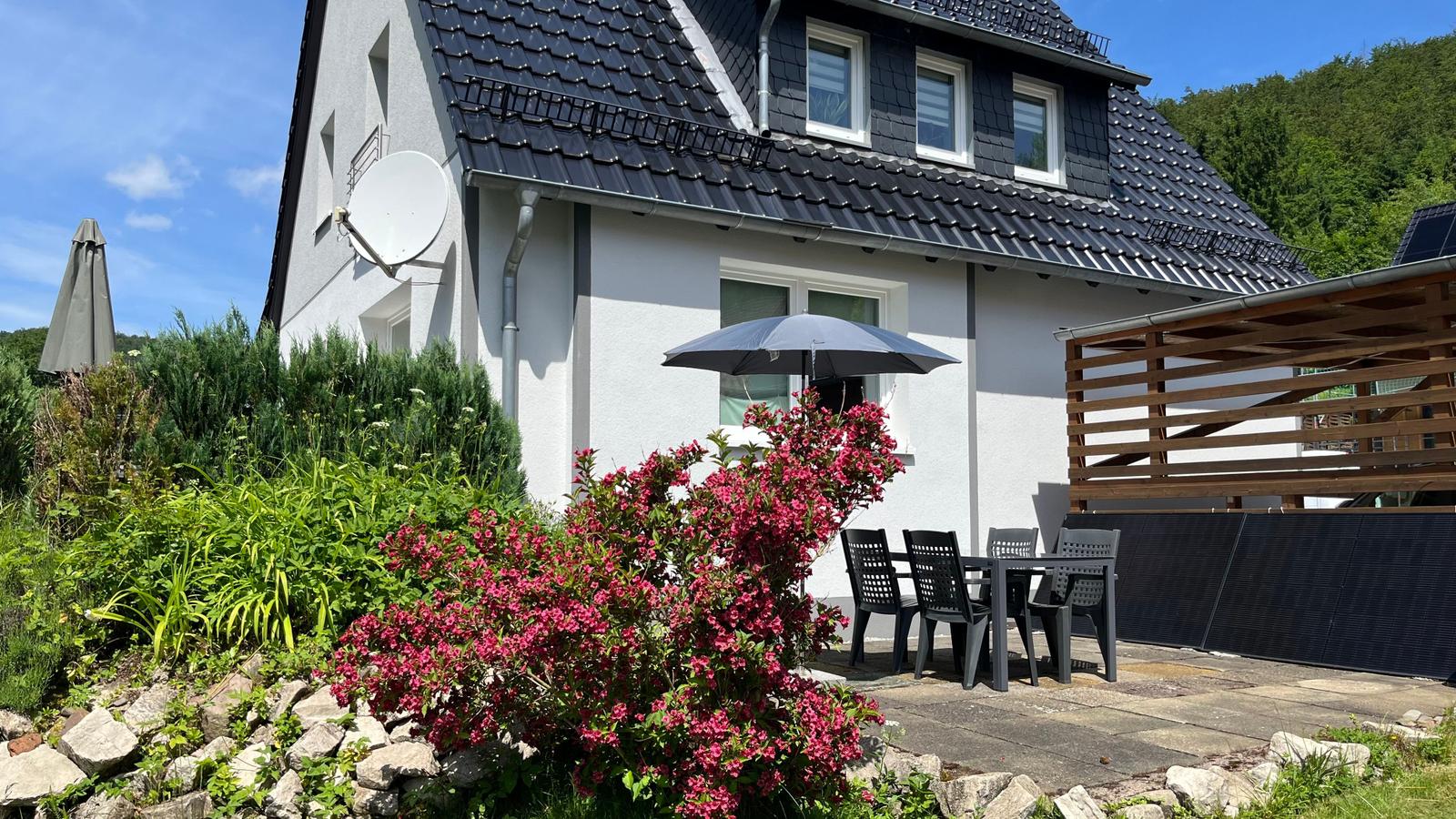 Haus mit Terrasse, Sitzgruppe und Blumenbeete. Schwarze Dachziegel und weiße Fassade.