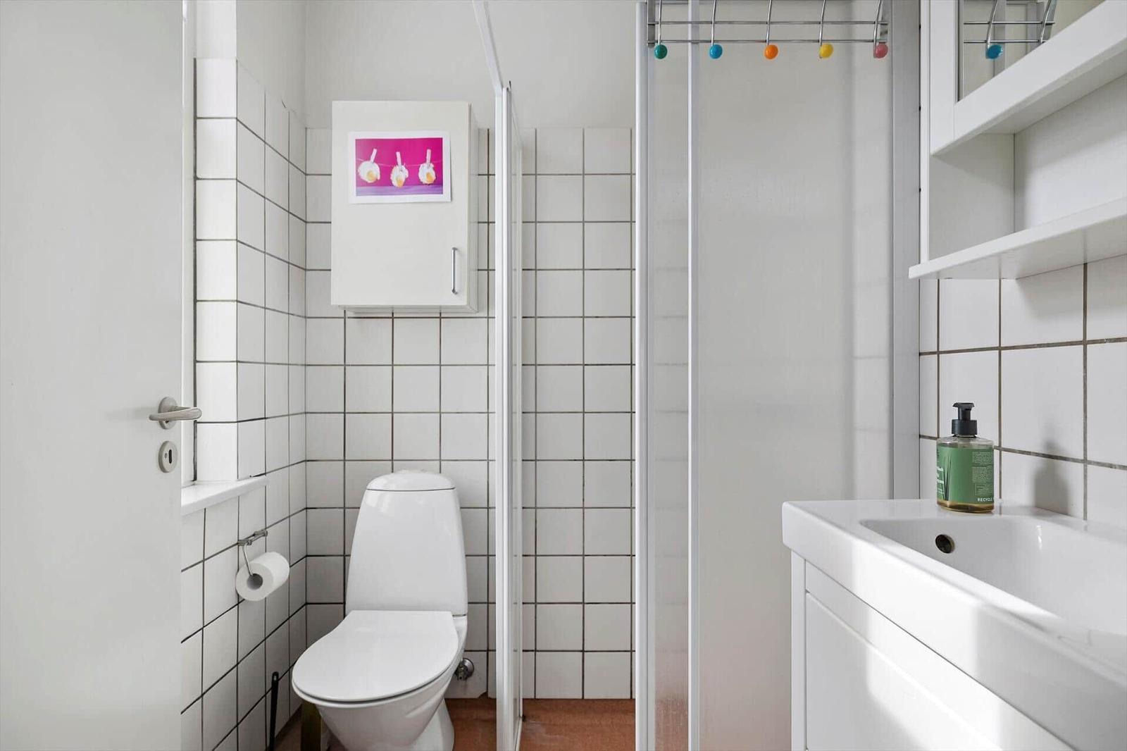 Bathroom with toilet, shower, and sink. White tiles and storage shelves.
