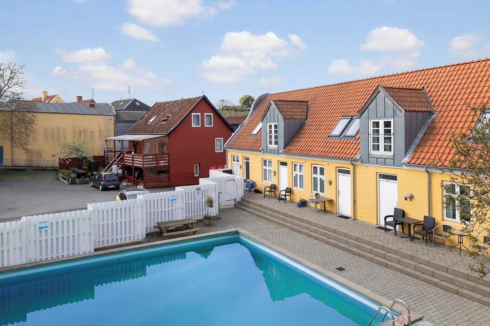 Pool area with yellow houses and white fence. Outdoor furniture and red roofs.