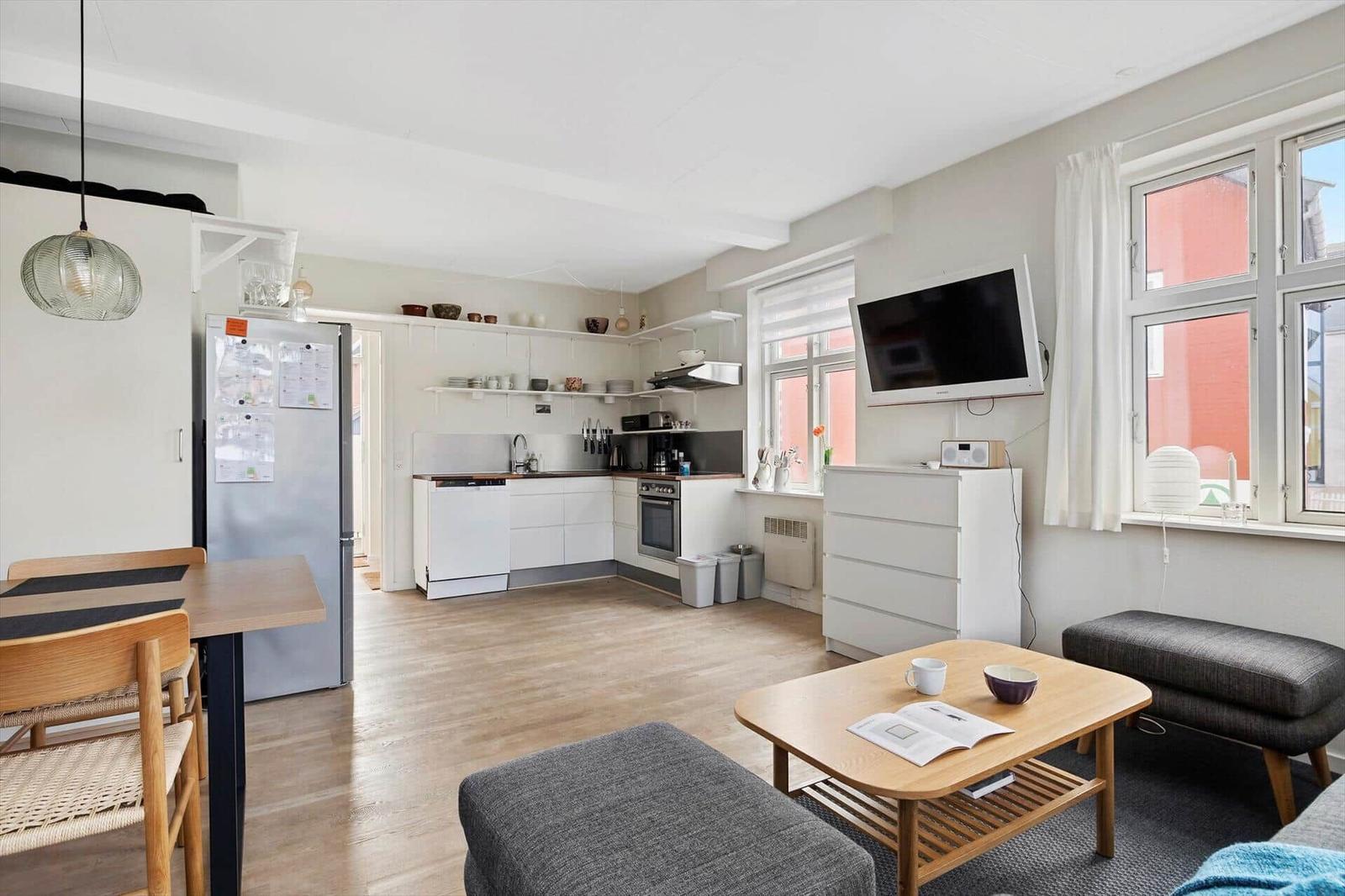 Living kitchen with dining table, fridge, and TV. Wooden floor and large windows.