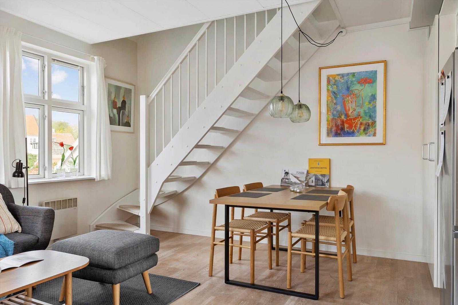 Dining area with wooden table and chairs under a staircase. Window, wall art, and refrigerator are visible.