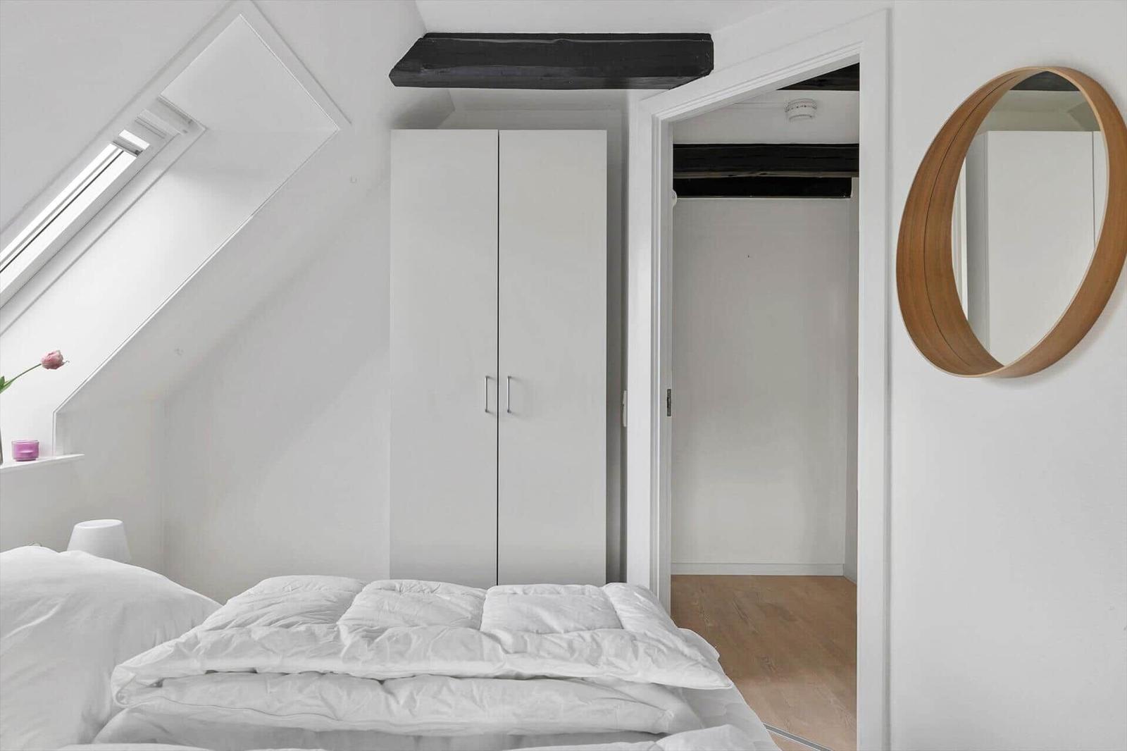 Bedroom with sloped ceiling, white bed, and wardrobe. Round mirror on the wall.