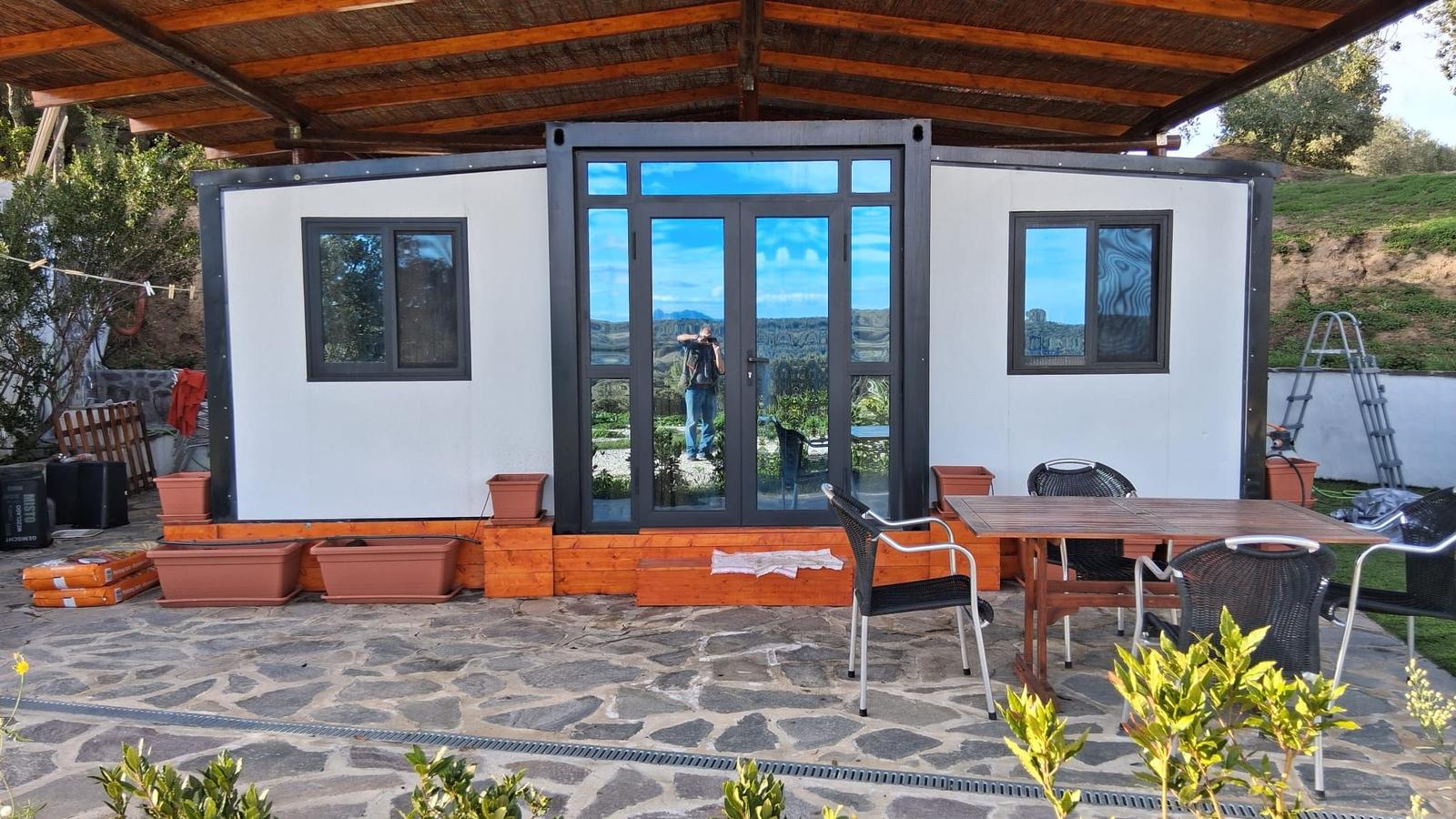 Container house with patio, table and chairs under wooden cover.