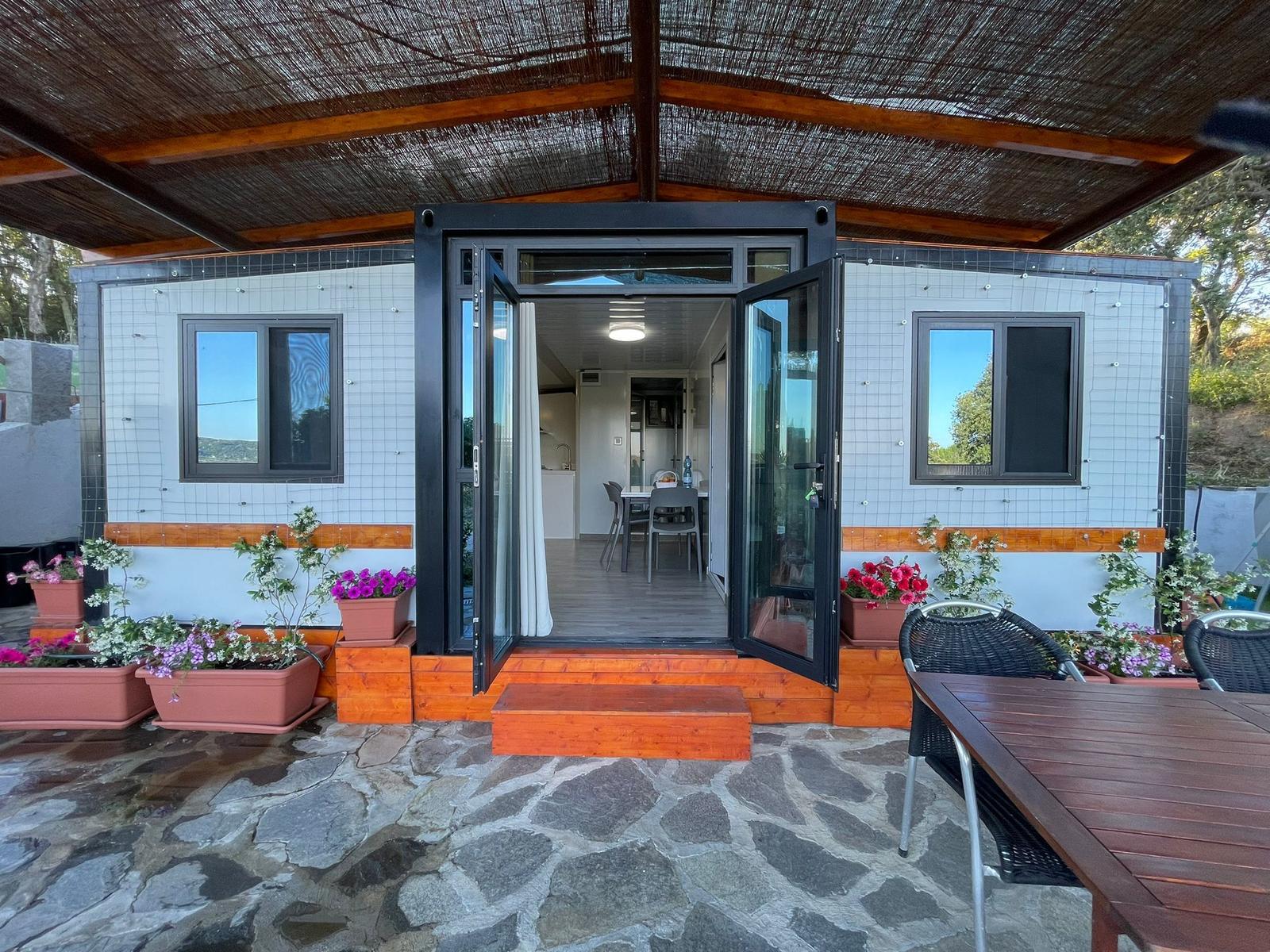 Container house with terrace, glass doors, and flower beds. Stone floor and wooden steps.