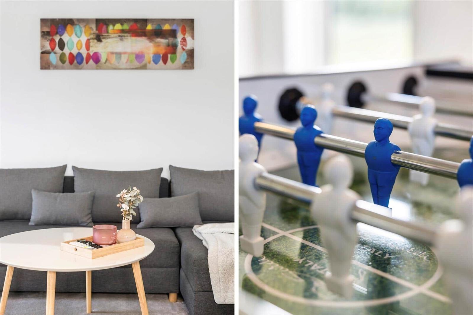 Graue Couch mit Kissen, runder Tisch und Wandbild. Foosballtisch mit blauen und weißen Figuren.
