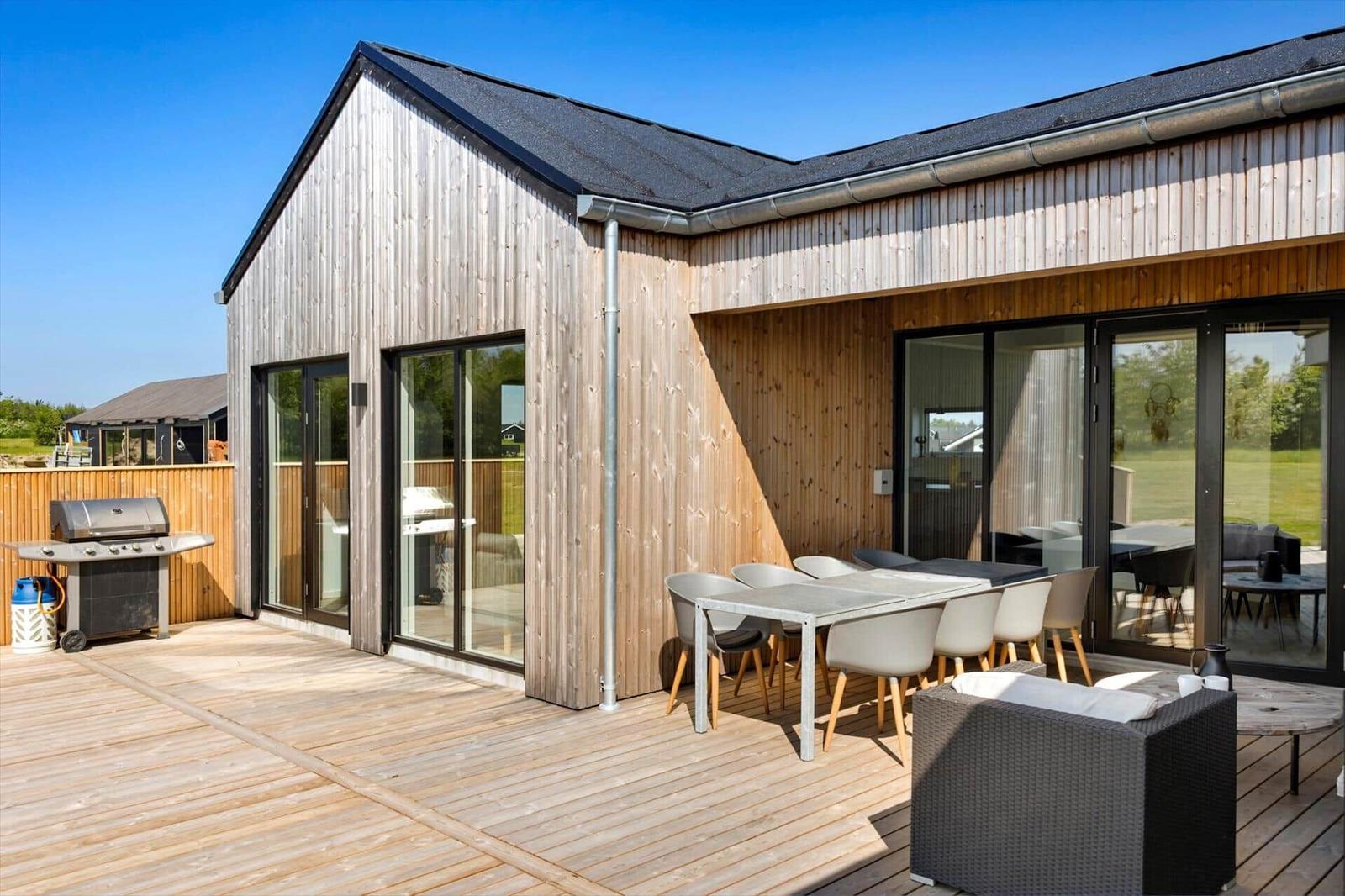Holzterrasse mit Grill, Esstisch und Sitzgelegenheiten. Glasfront führt ins Haus.