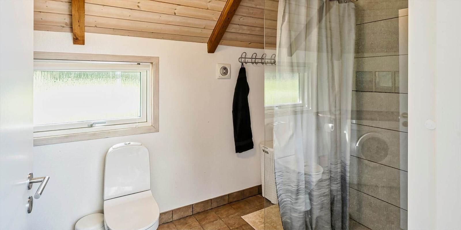 Badezimmer mit Toilette, Dusche und Fenster. Holzdecke und Badewanne sichtbar.