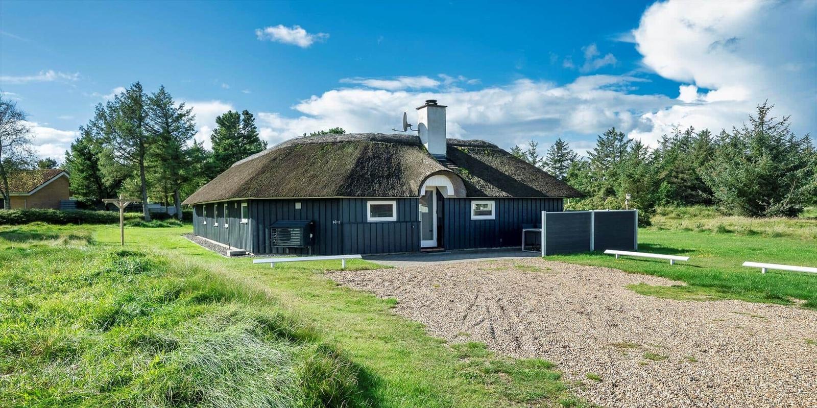 Dorfhaus mit Strohdach, Eingang und Garten. Umgeben von Bäumen und Gras.
