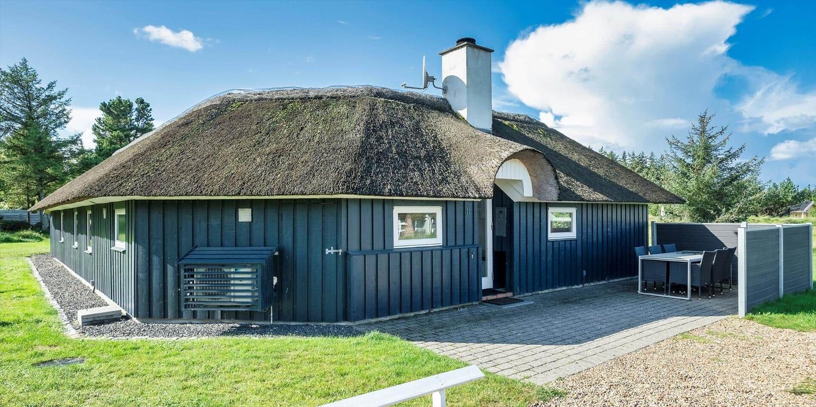 Ein Haus mit strohgedecktem Dach und dunkelblauer Fassade. Außenbereich mit Tisch und Stühlen.