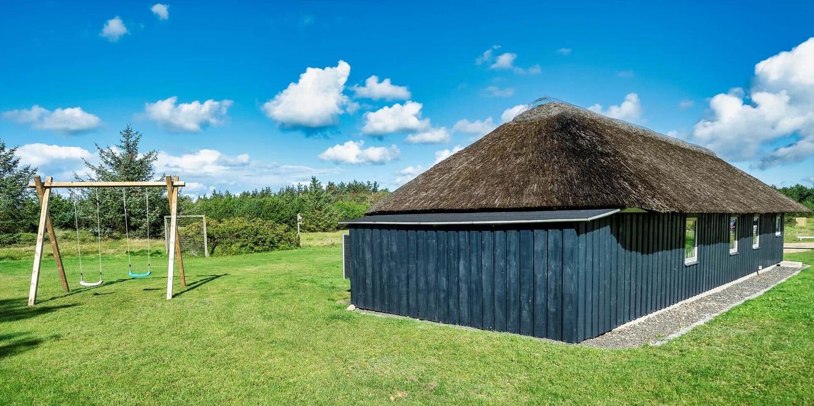 Ein Haus mit Strohdach und dunkler Holzfassade auf einer grünen Wiese mit Schaukel.