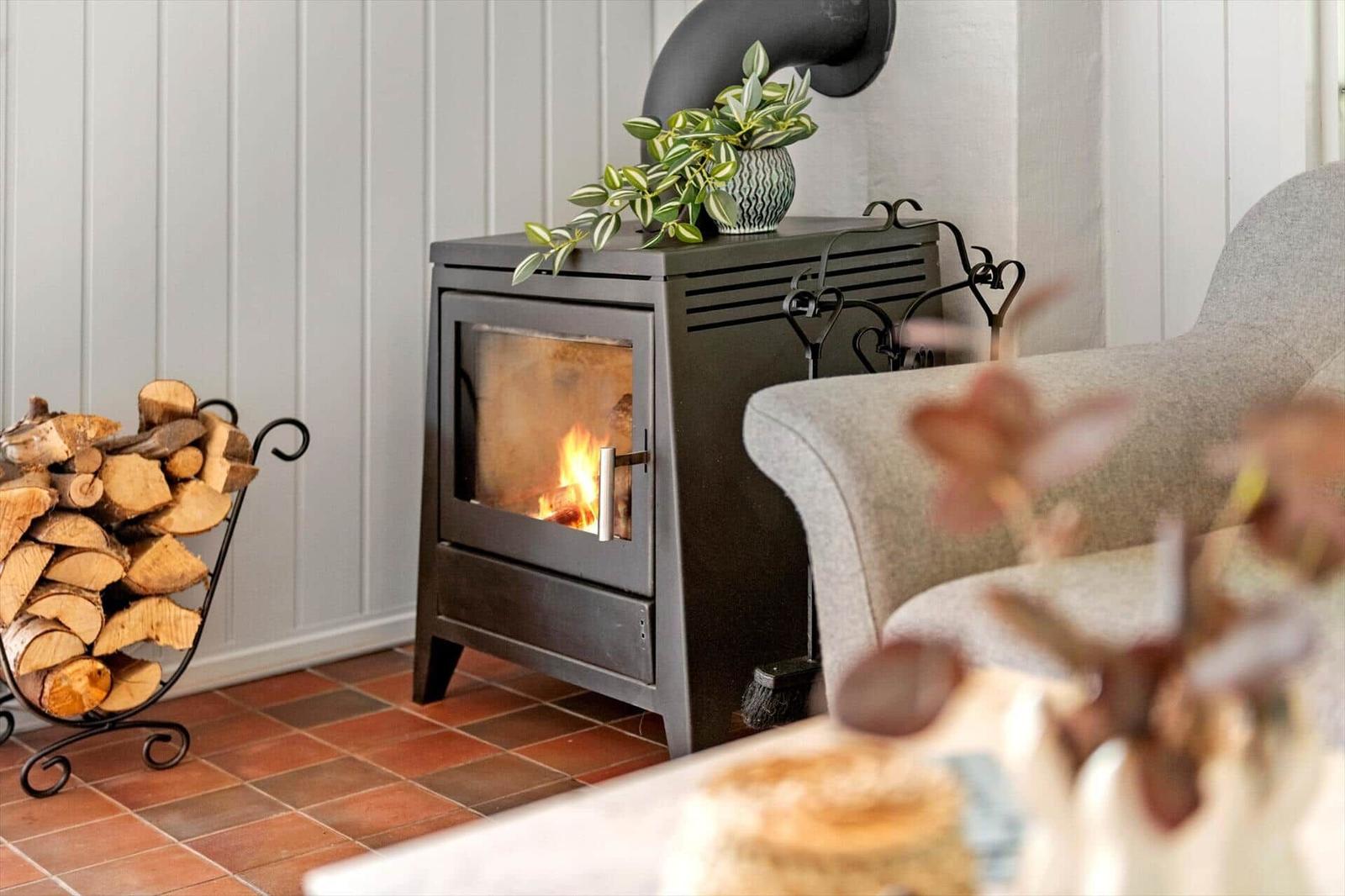 Ein Kaminofen mit brennendem Feuer, daneben ein Holzstapel und ein grauer Sessel.