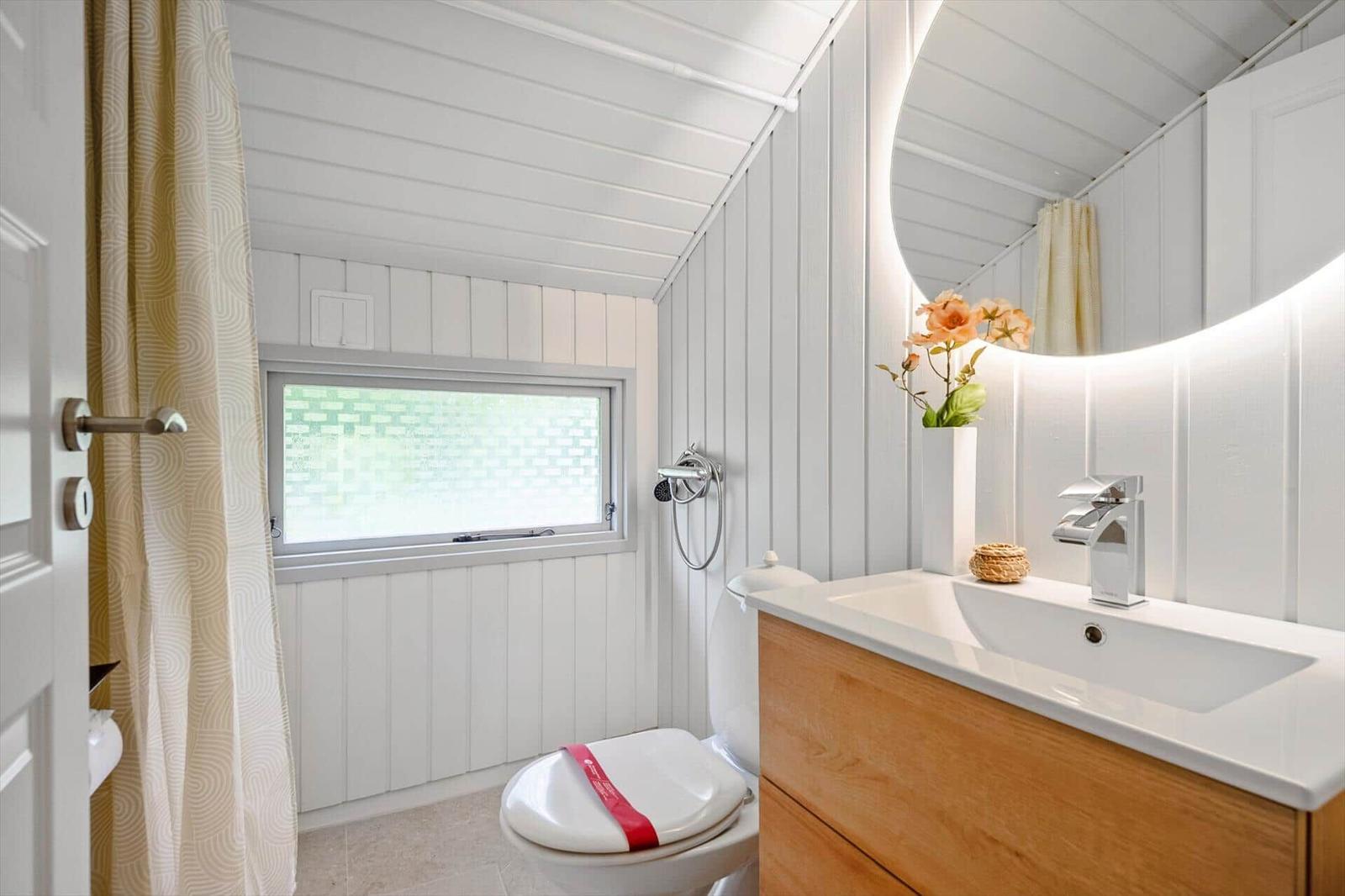 Badezimmer mit Toilette, Waschbecken und Dusche. Weiße Wände, Holzschrank und Fenster.