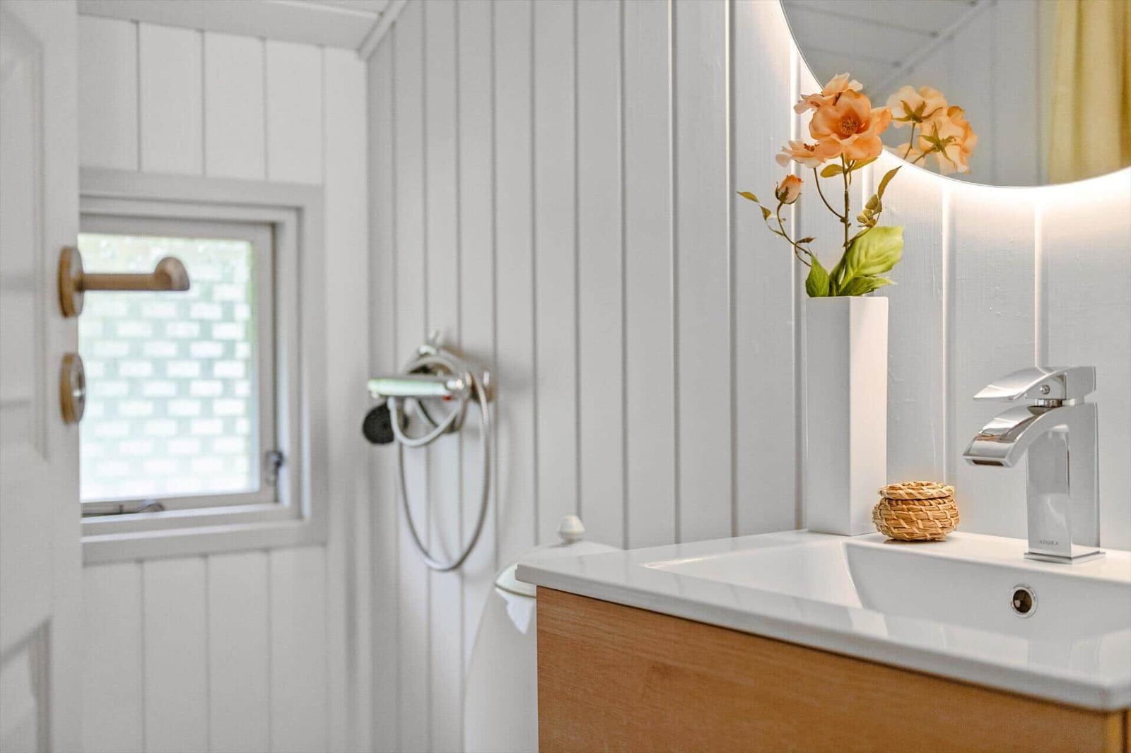 Badezimmer mit Waschbecken, Spiegel und Dusche. Holzunterschrank und Blumen im Vasen.