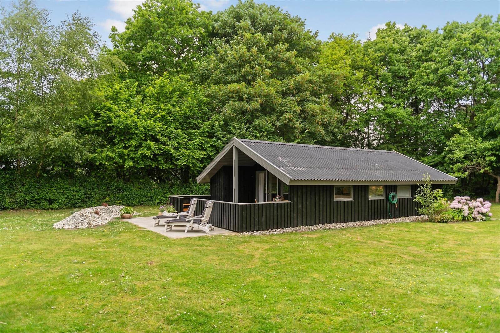 Schwarze Hütte mit Terrasse, Liegestühlen und grünem Garten hinter Bäumen.