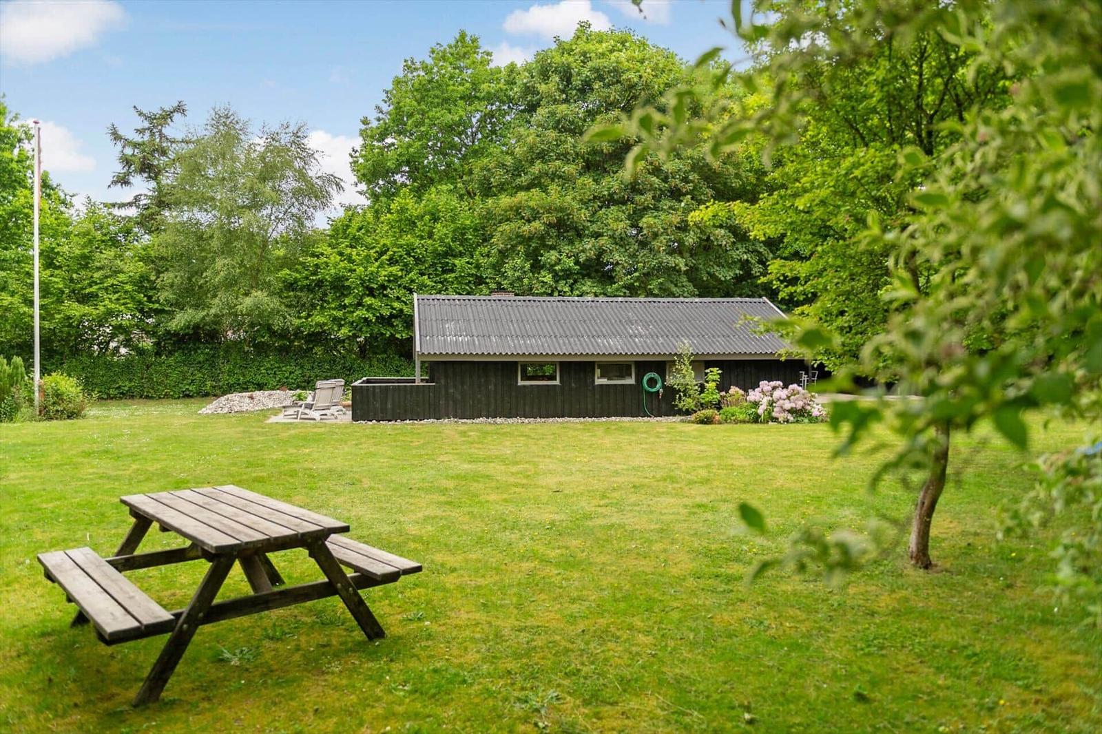 Hütte mit Garten, Picknicktisch und Blumenbeet. Umgeben von Bäumen und Gras.