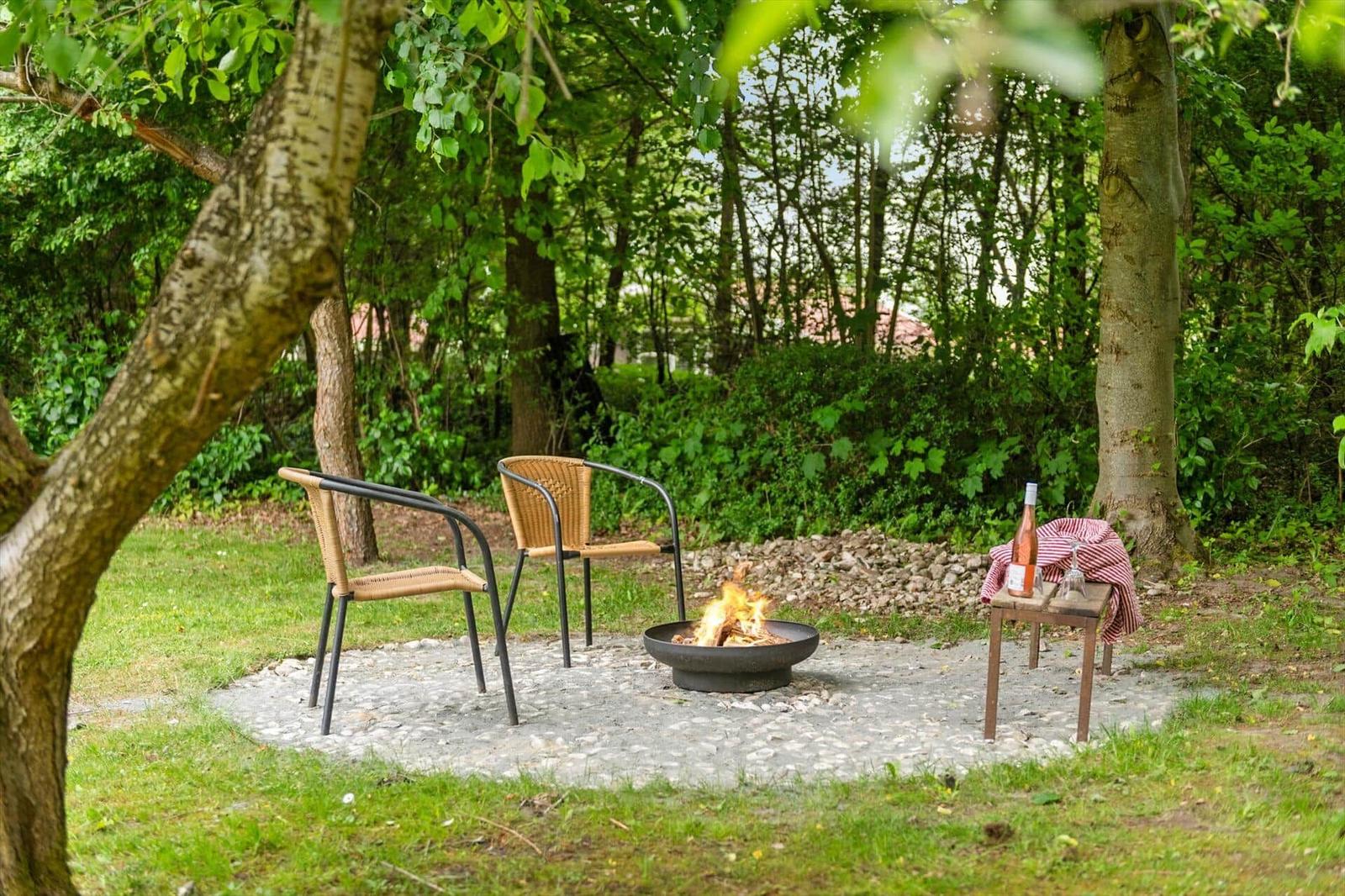 Feuerkorb mit zwei Stühlen im Grünen. Tisch mit Weinflasche und Glas.