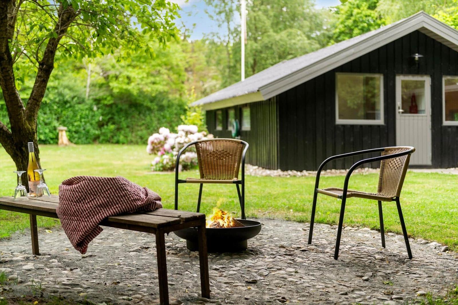 Außenbereich mit Feuerkorb, zwei Stühlen und Tisch unter Bäumen vor einem Haus.