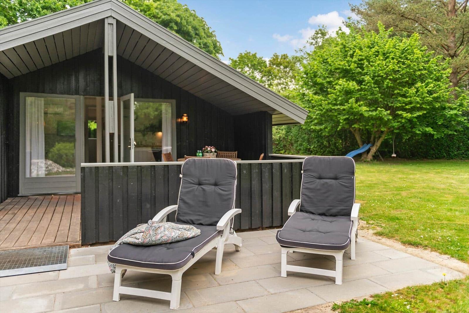 Terrasse mit zwei Liegestühlen vor einem Holzhaus mit Garten und Bäumen.