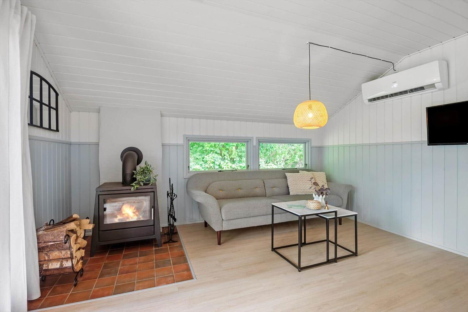 Wohnraum mit Sofa, Kaminofen und Fenstern zum Grün. Decke und Wände sind weiß.