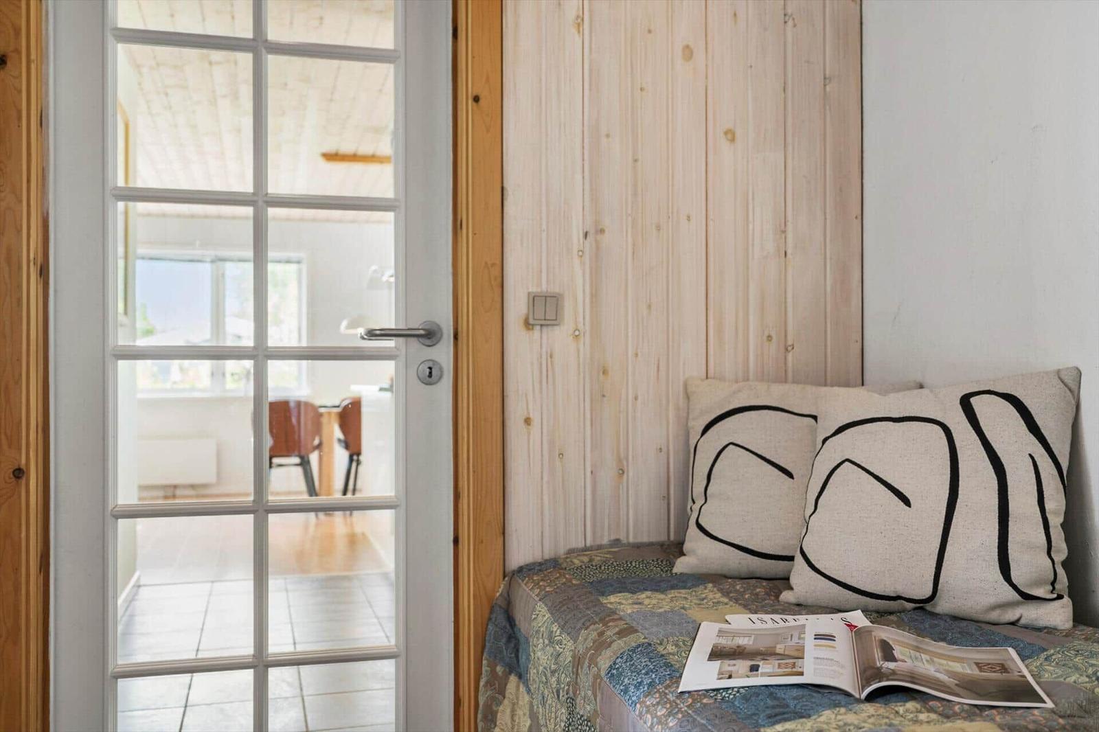 Schlafzimmer mit Holzwand und Tür zum Wohnbereich. Auf dem Bett liegen Kissen und ein Magazin.