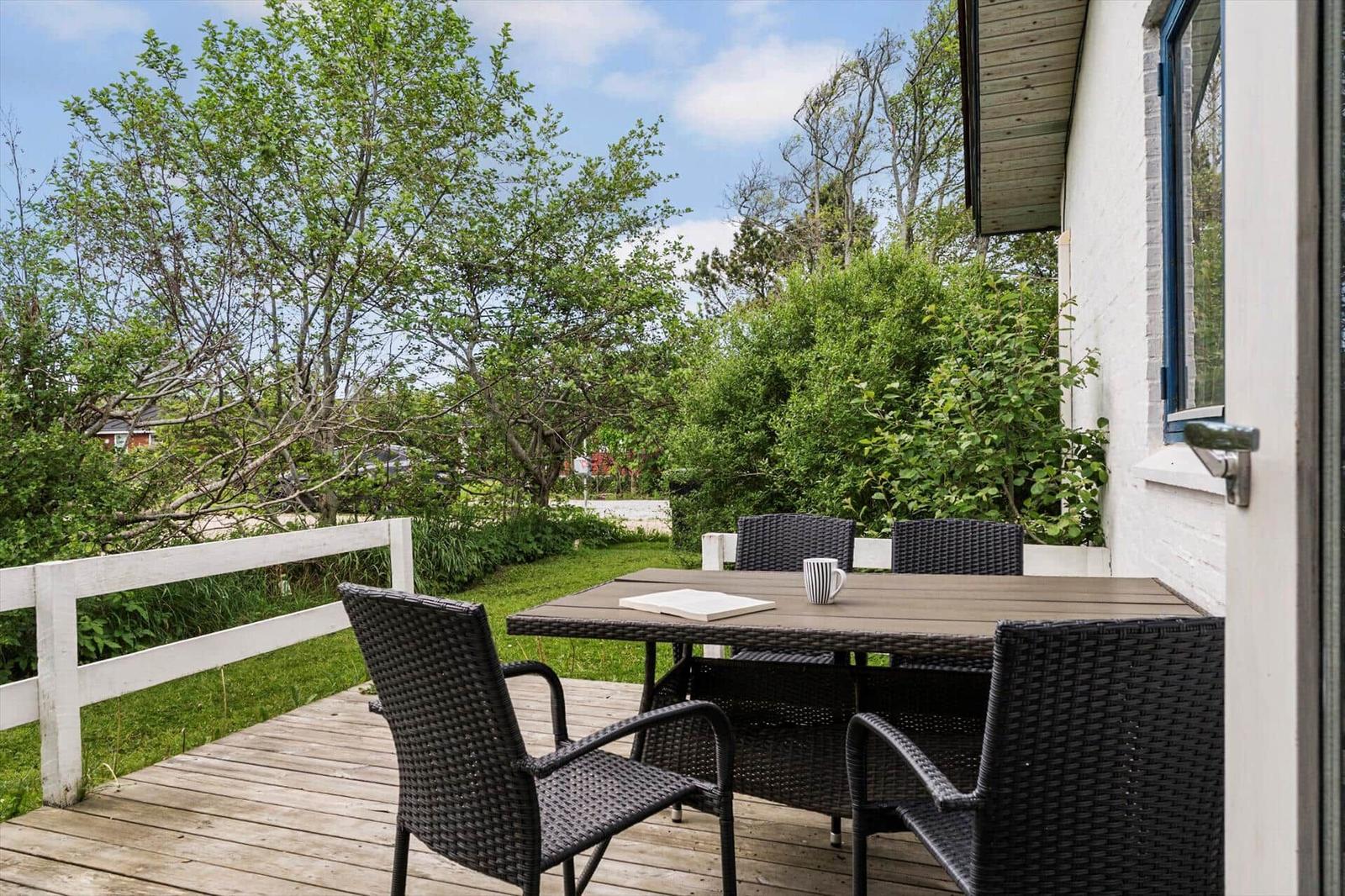 Terrasse mit Tisch und Stühlen, umgeben von Grün und einem weißen Zaun.
