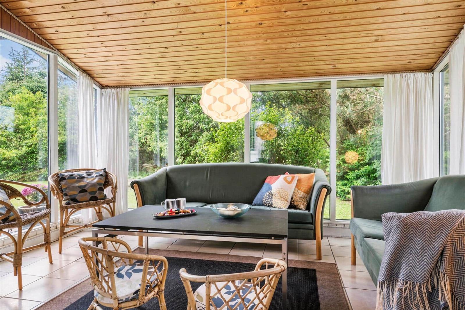 Wohnbereich mit Sofa, Stühlen und Tisch. Holzdecke und große Fenster mit Blick auf Garten.