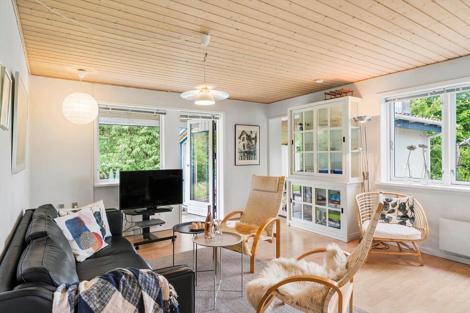 Wohnzimmer mit Sofa, Sessel und TV. Holzdecke und große Fenster.