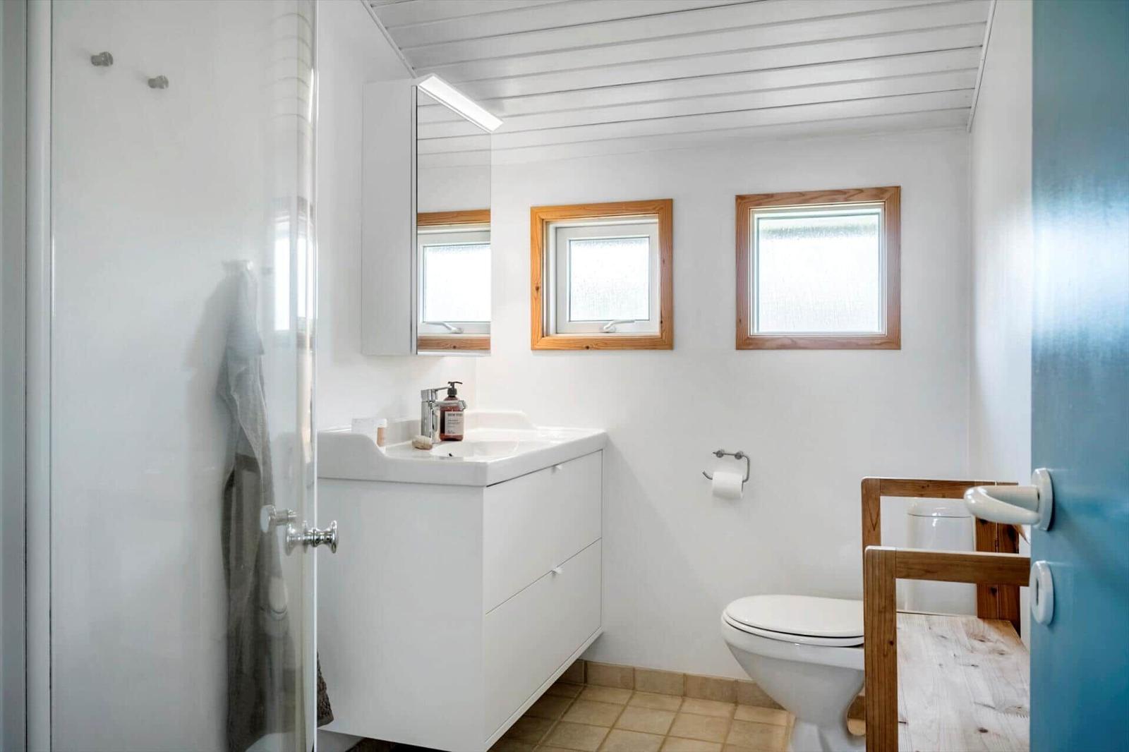 Badezimmer mit Dusche, Waschbecken und Toilette. Zwei Fenster mit Holzrahmen.