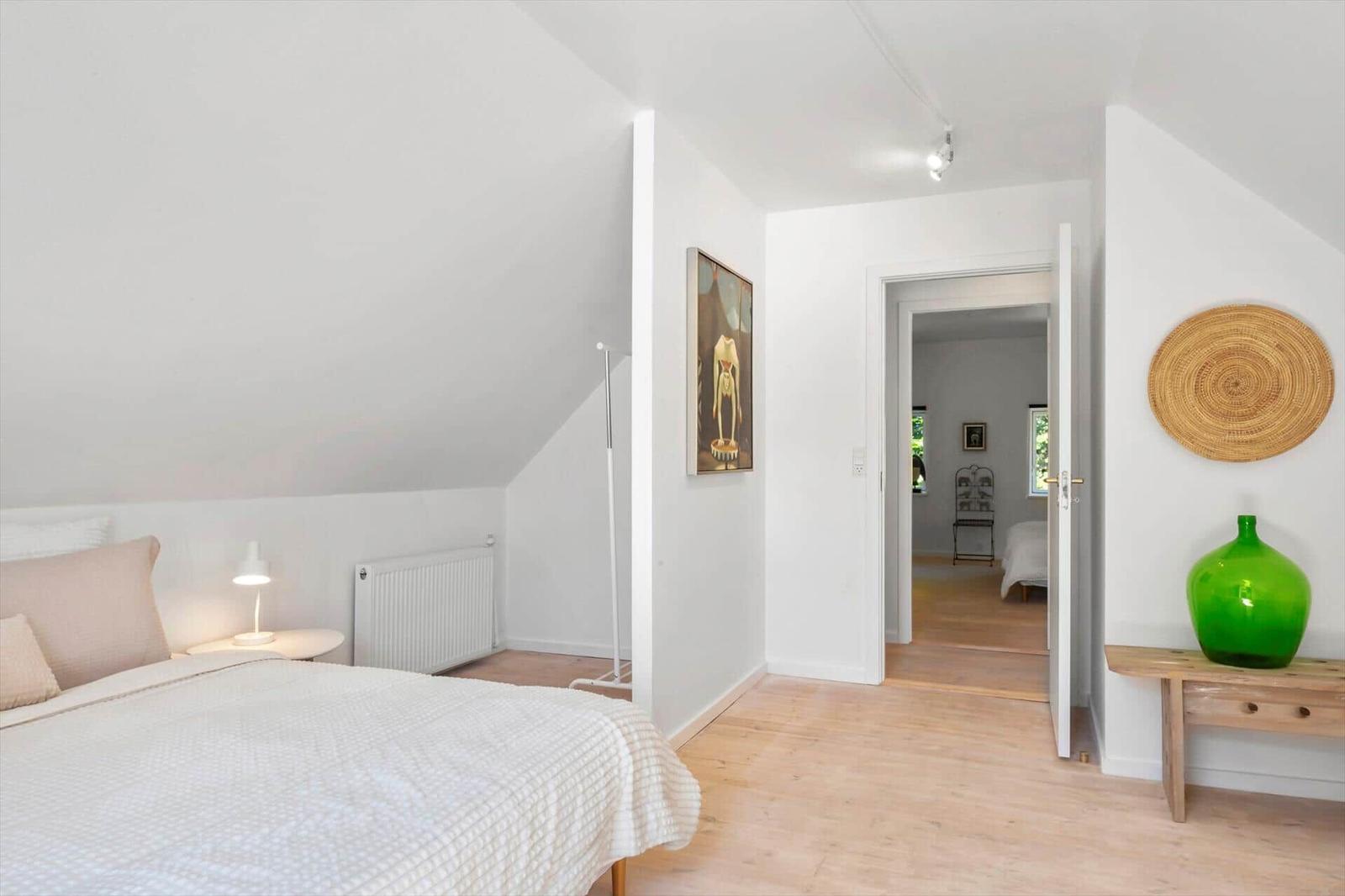 Schlafzimmer mit Bett, Nachttisch und grüner Vase. Tür führt in angrenzenden Raum.