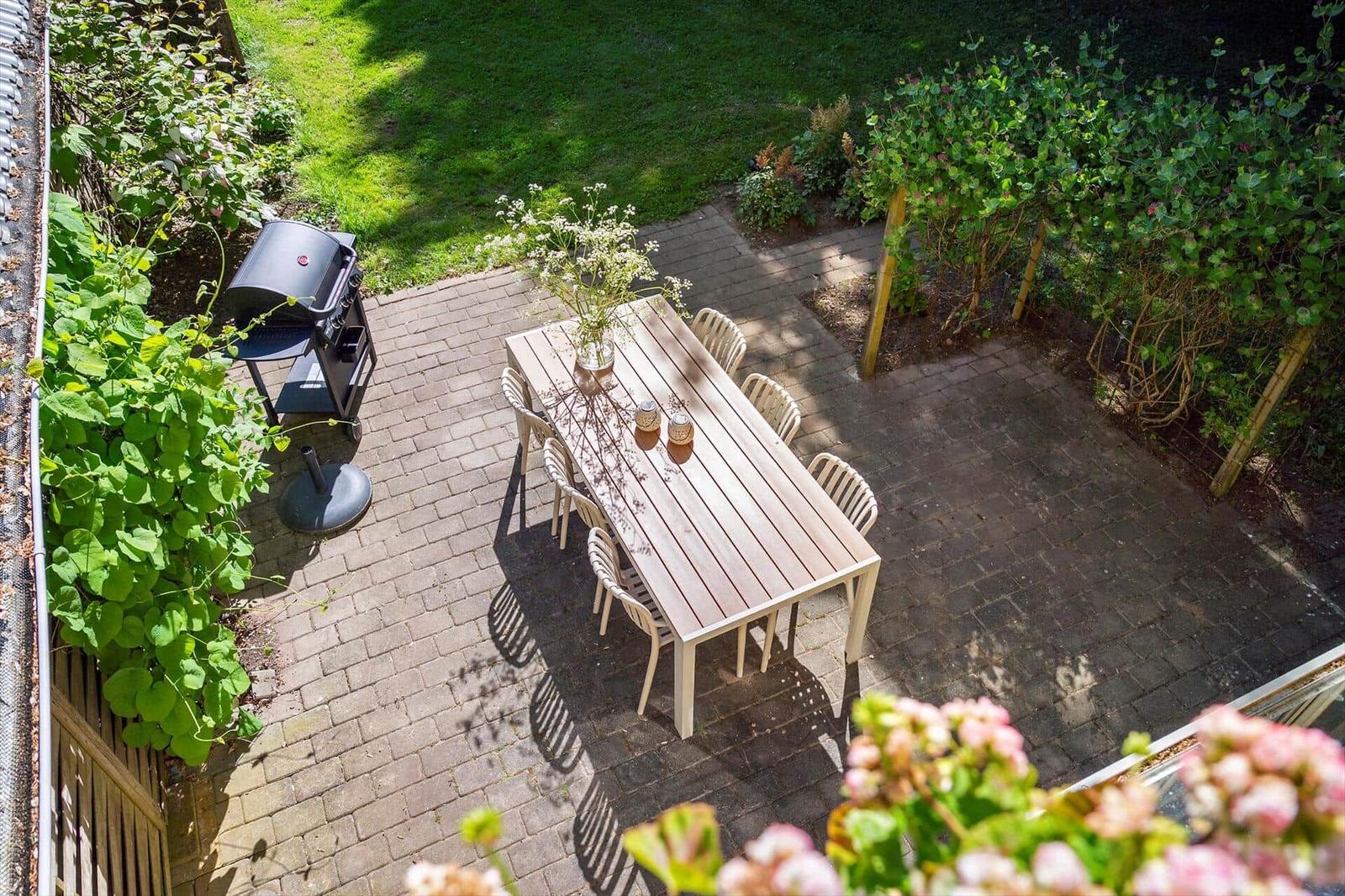 Terrasse mit Holztisch, sechs Stühlen, Grill und Blumenbeeten.