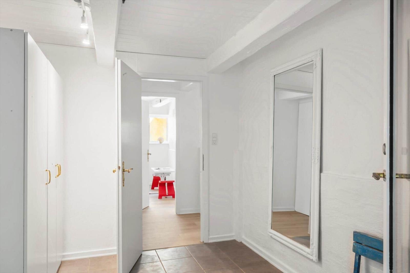 Flur mit Spiegel, Tür und Kleiderschrank. Durchgang zu einem Raum mit Waschbecken und roten Hocker.