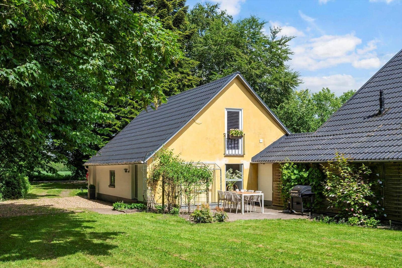 Gelbes Haus mit Terrasse, Grill und Garten unter Bäumen.