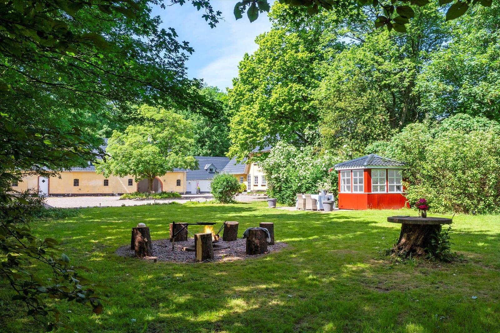 Grüner Garten mit Feuerstelle, Holzstühlen und rotem Pavillon. Im Hintergrund sind Gebäude und Bäume zu sehen.