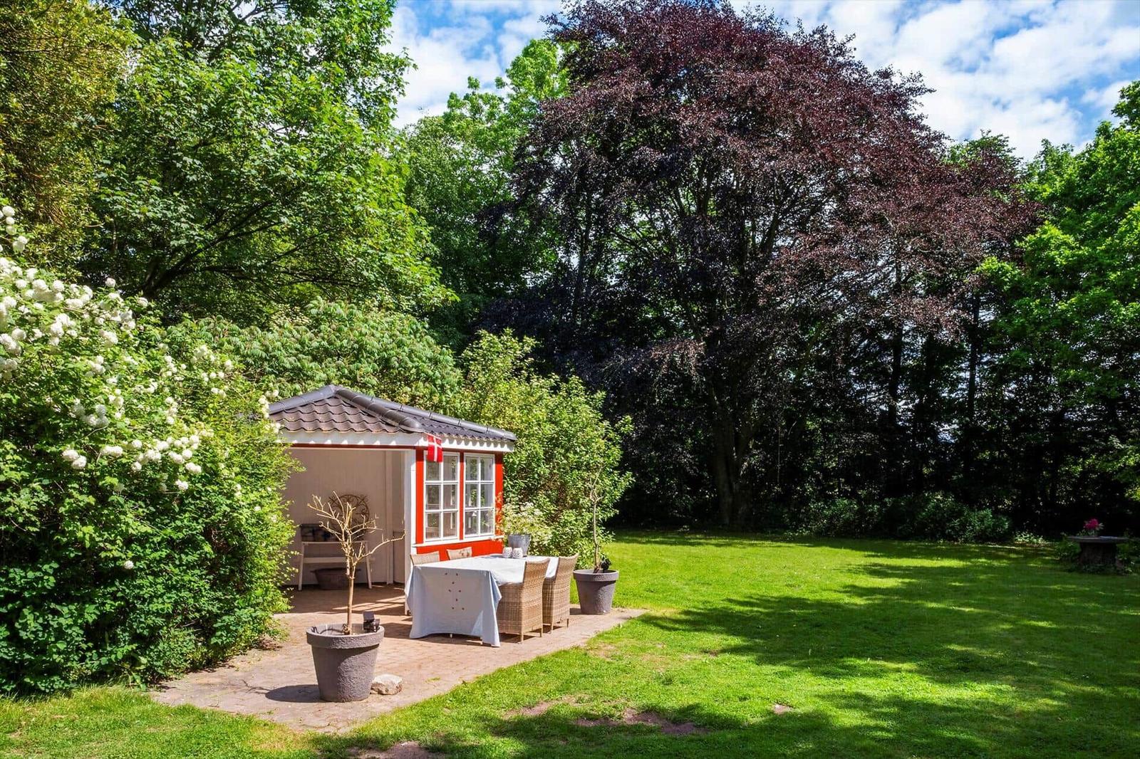 Ein kleiner Pavillon mit Tisch und Stühlen im Garten. Umgeben von Bäumen und Gras.