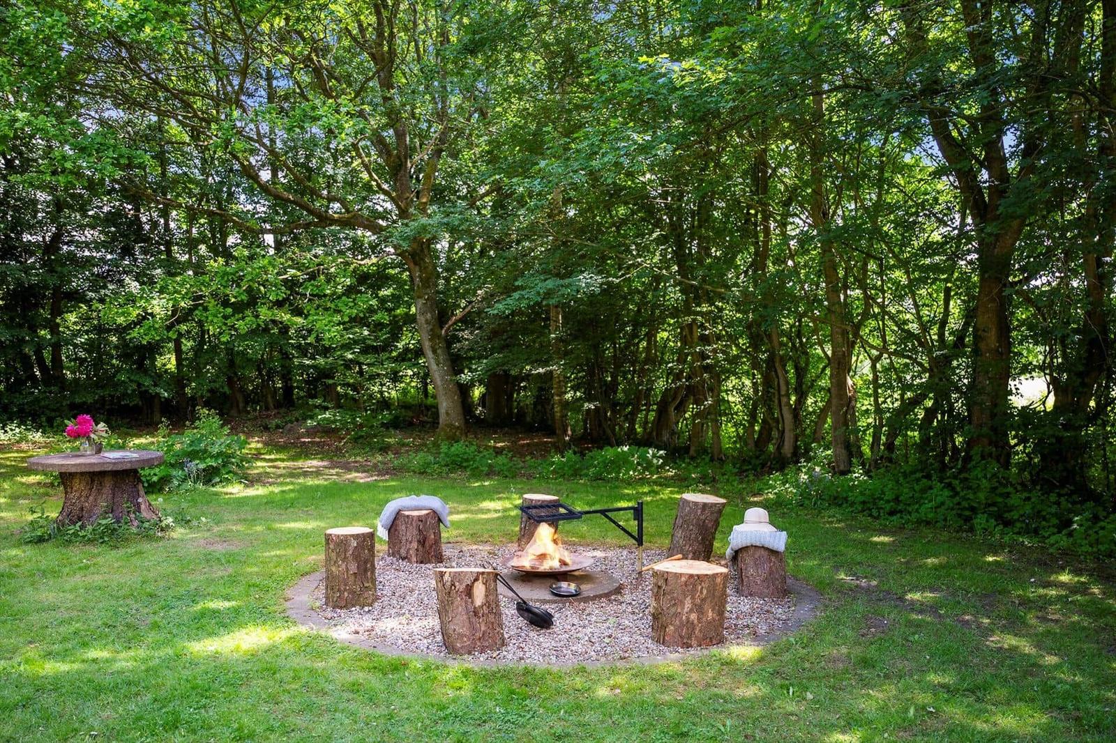 Feuerplatz mit Holzstümpfen und Grill im Wald. Tisch mit Blumen daneben.