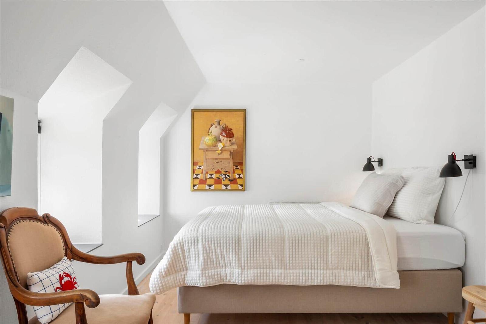 Schlafzimmer mit Bett, Stuhl und Wandbild unter schrägen Dachdecken.