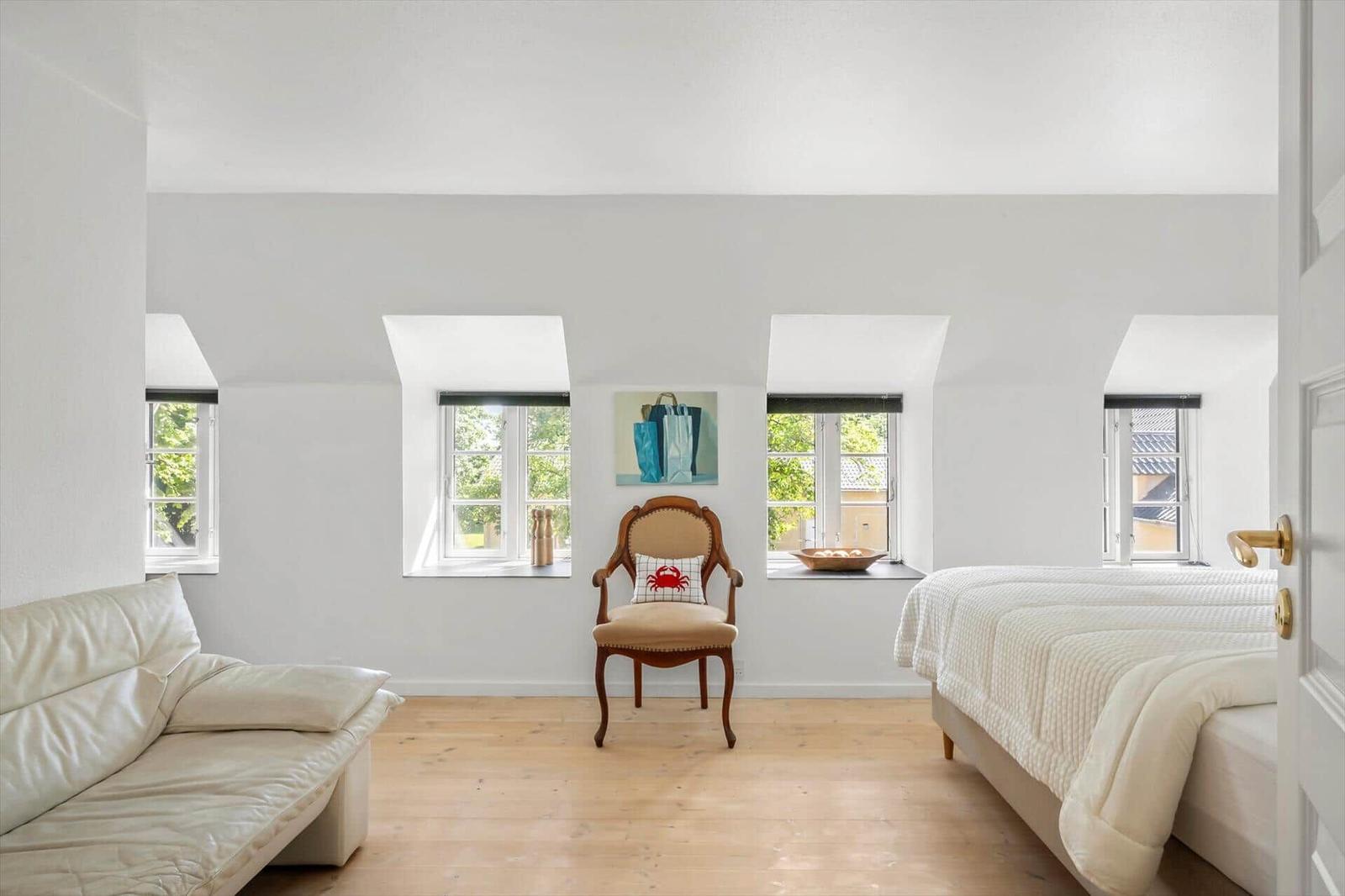 Ein Schlafzimmer mit Bett, Sofa und Stuhl. Vier Fenster und helles Holzfußboden.