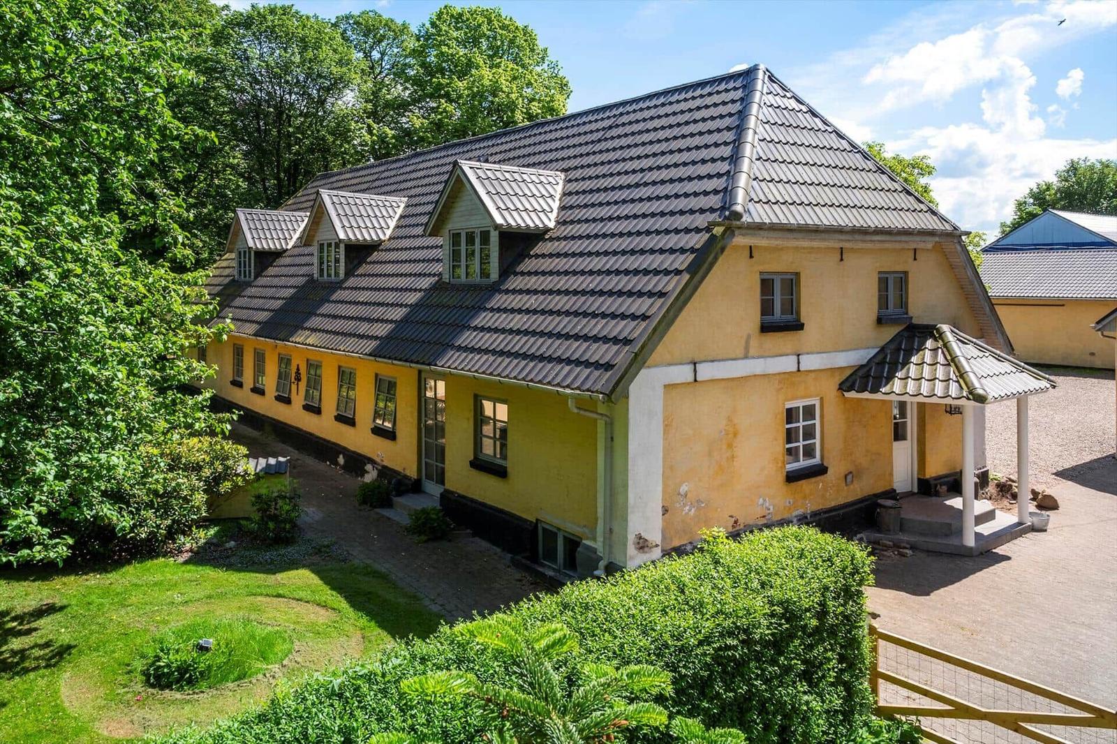 Ein gelbes Haus mit dunklem Dach, umgeben von Grün und einem gepflasterten Hof.