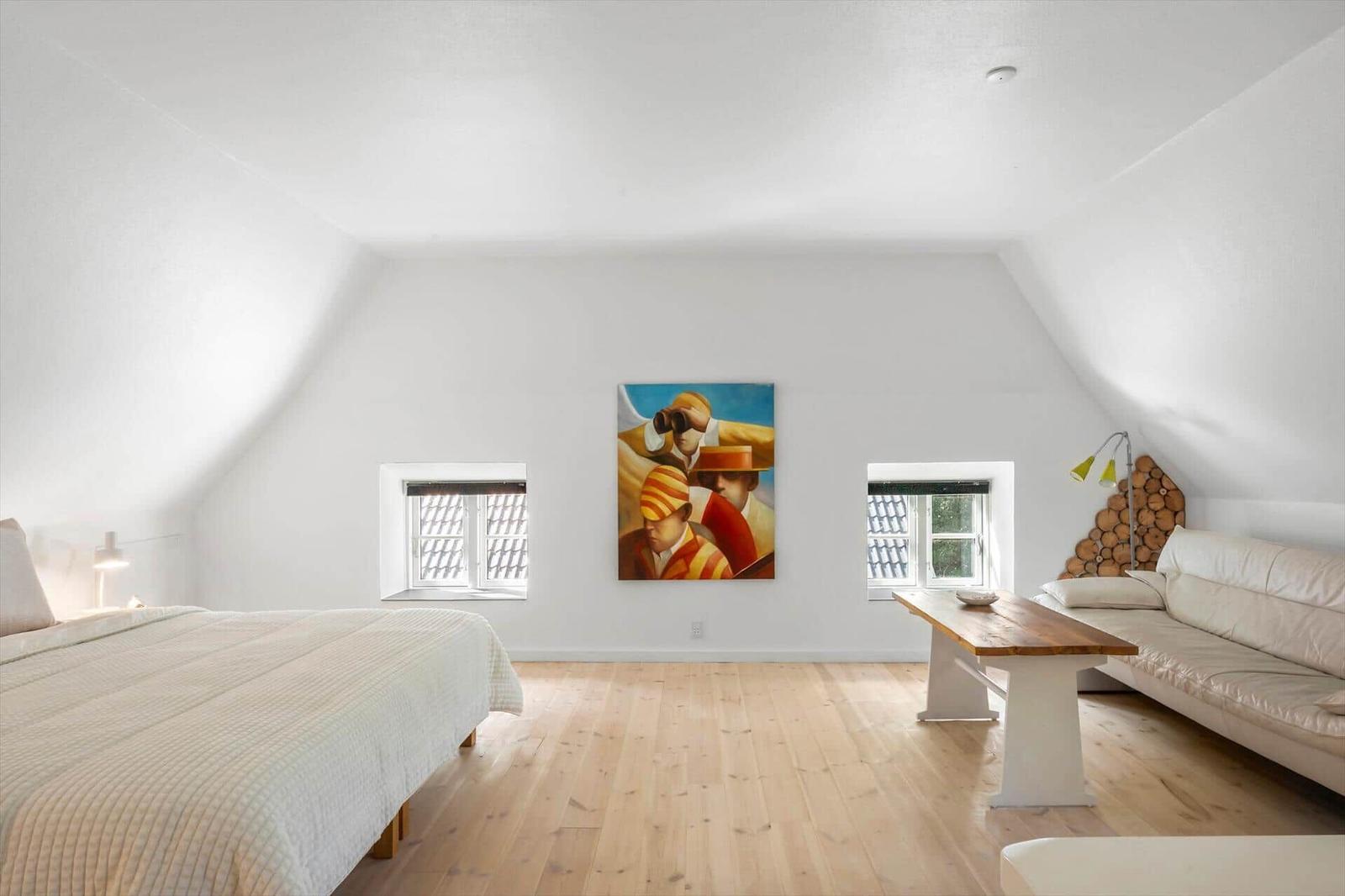 Ein Raum mit Bett, Sofa, Tisch und Wandbild zwischen zwei Fenstern.