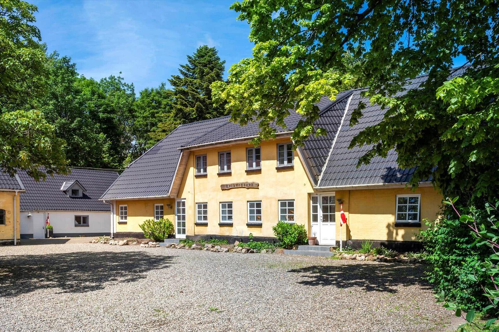 Ein gelbes Haus mit grauem Dach und weißen Fenstern, umgeben von Bäumen und einem Kiesplatz.