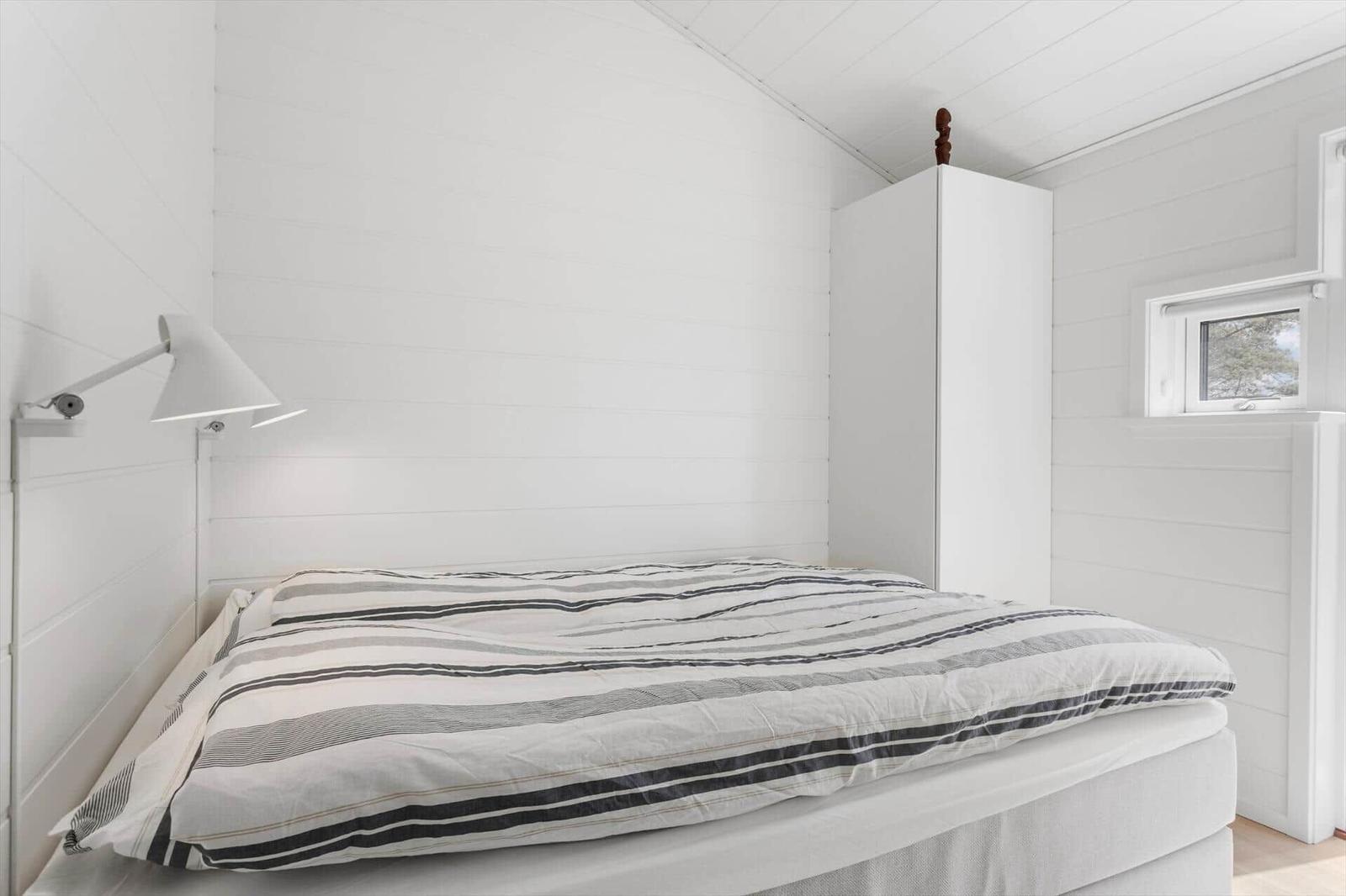 Schlafzimmer mit weißer Wandverkleidung, Bett mit Streifenbezug und Fenster.