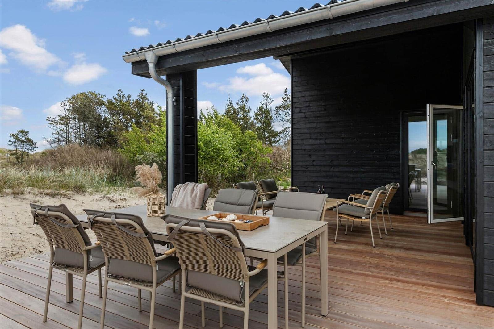 Terrasse mit Tisch und Stühlen, Blick auf Dünen und Bäume.