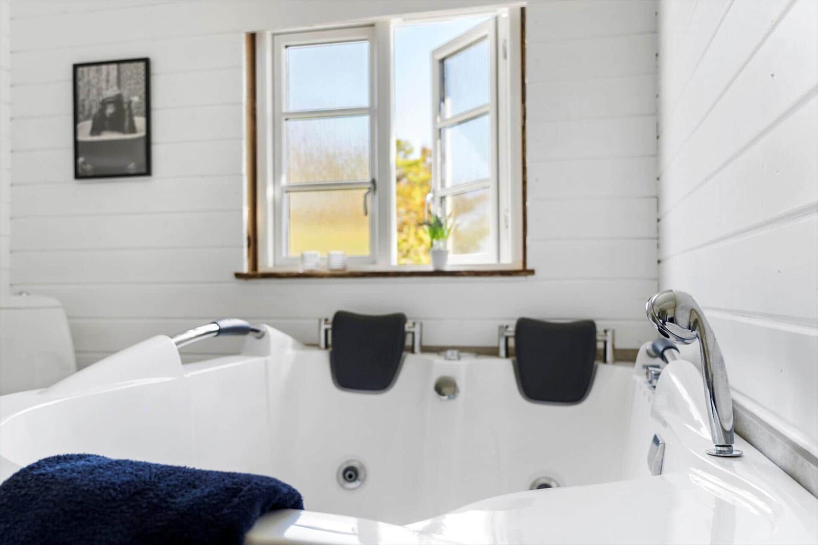 Eine weiße Badewanne mit schwarzen Kissen und einem blauen Handtuch. Ein Fenster zeigt eine Außenszene.