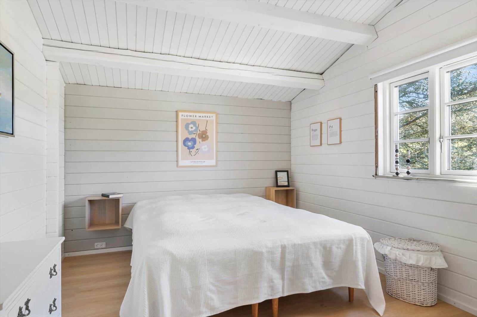 Schlafzimmer mit weißem Bett, Holzdielen und Fenster mit Blick auf Bäume.